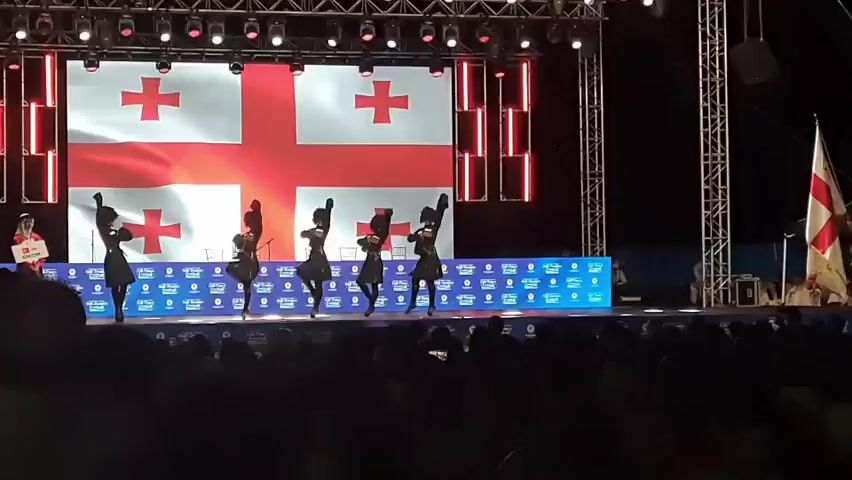 Five young individuals dressed in traditional Georgian dance attire are performing on a stage. They are all wearing black tops and skirts with white accents. Their hair is styled in dark, curly wigs. The backdrop of the stage features a large projection of the Georgian flag, which consists of a white background with a large red cross and four smaller red crosses in each of the four quadrants. The stage is lit with bright stage lights, and there are vertical red LED lights on either side of the flag. In front of the stage, a blue barrier with multiple logos is visible. A crowd of spectators is seated in the foreground, their faces largely obscured by darkness. To the far left of the stage, a person holding a white sign with red writing can be seen. To the far right, a flagpole with a white flag and a red cross is visible. The performance appears to be part of a larger event or competition.