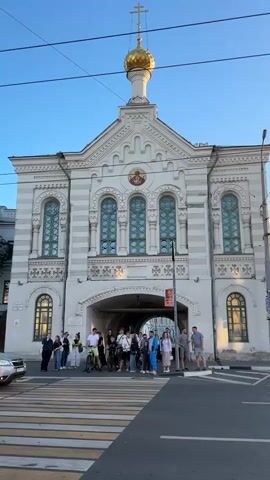 The video shows a street scene with a white building featuring a golden dome topped with a cross. The building has arched windows and decorative elements. In front of the building, a group of people, some on bicycles, are gathered near a crosswalk. A silver SUV is driving across the crosswalk, and a black car is also visible. The sky is clear and blue.