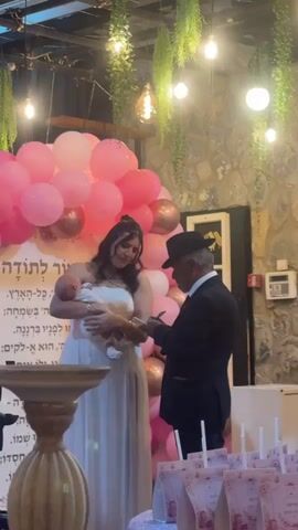 A woman in a white dress holds a baby while standing next to an older man in a black suit and hat. They are surrounded by pink balloons and greenery, with a stone wall in the background. The man appears to be writing something on a piece of paper. A young woman in a red dress is visible in the background.