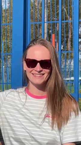 A fair-skinned woman with light brown hair stands facing the camera. She is wearing sunglasses, a light-colored, striped t-shirt with a pink collar, and a logo on the front. She is smiling, and is briefly covered with soap bubbles that fly into the frame. Behind her is a tall blue metal fence and trees. The scene appears to be outdoors in the daytime.