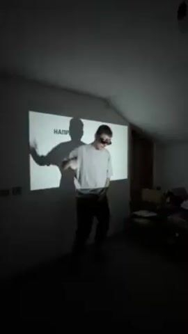 The video shows a young man dancing in front of a projector screen. The room appears to be an attic with dark floors and white walls. The man is wearing black pants, a light grey T-shirt, and dark sunglasses. The projector screen displays large black text in a font that resembles block letters. The man's shadow is also visible on the screen behind him. He appears to be enjoying himself as he moves to the music.