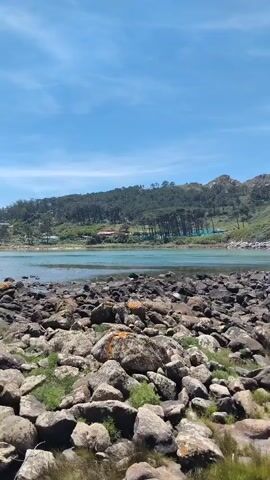 This video shows a rocky shoreline with a body of water. In the background, there are hills covered with trees. A narrow, stone walkway or pier extends into the water. The sky is blue with some scattered clouds. The sun is shining, creating bright lighting. The video pans slightly from left to right, showcasing the landscape. There are no people or animals visible in the video.