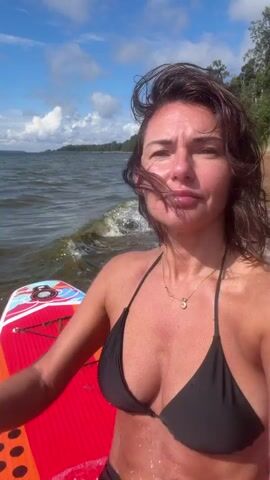 The video shows a woman in a black bikini top and black leggings on a paddleboard in the water. She is taking a selfie video. The water is choppy, and there is a beach in the background. The sky is blue with some clouds. The woman is moving her head and body, and the paddleboard is also moving in the water. She makes various facial expressions, including smiling and pouting.