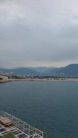 The video showcases a coastal landscape under a cloudy sky. The deep blue sea dominates the foreground and mid-ground, appearing calm. In the background, majestic mountains line the coast, with a town or settlement visible at their base. Green foliage, including bushes, small trees, and palm-like plants, adorns the cliffside in the foreground, suggesting an elevated viewpoint.

At one point, a woman wearing a black t-shirt with a colorful abstract design and glasses is seen holding up a smartphone, seemingly capturing the scenic view. She has a white shoulder bag with metallic accents. Behind her, a wooden pole and a modern white structure (possibly part of a building or gazebo) are visible. The overall atmosphere is peaceful, with overcast conditions suggesting a mild day. No explicit text is visible in the video. The setting aligns with a coastal area like Finike, Turkey.