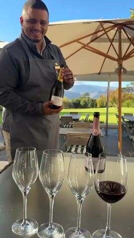 The video shows a man dressed in a grey uniform pouring sparkling wine into three stemmed wine glasses arranged on a table outside. To the right, another bottle of sparkling wine is visible, along with a wine glass containing red wine. The man is focused on pouring the sparkling wine. The background reveals a scenic landscape with green grass, trees, and mountains in the distance, all under a bright sky. A patio umbrella is visible overhead.
