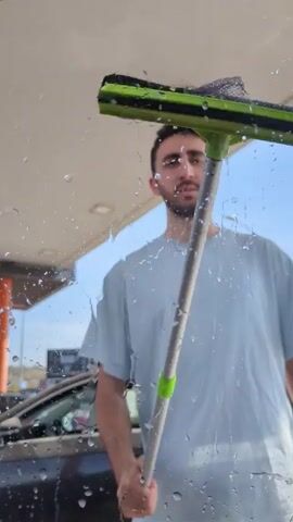 A man is seen through a wet glass as he cleans the window with a green squeegee. He is wearing a light blue t-shirt and has a beard. The man appears to be at a gas station. The sky is visible in the background.