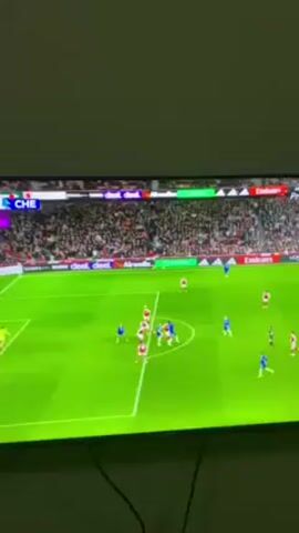 The video shows a football game being played on a large television screen. The camera is held in a portrait orientation, capturing the screen from below. The game appears to be between Arsenal (in red and white) and Chelsea (in blue). The game is at the 95th minute, and the score is Arsenal 2 and Chelsea 1. A player wearing a blue uniform is lying in the goal. The area around the goal is packed with players from both teams. The stadium is filled with a large crowd of fans. The television screen has several advertisements along the bottom and sides. A black cable is seen dangling in front of the TV. The wall in the background is beige. The overall lighting suggests the room is dim.