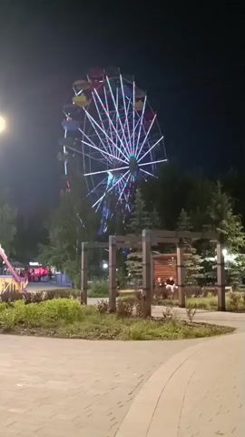 A Ferris wheel is illuminated with colorful lights against a dark sky. In the foreground, there is a paved area and some greenery. Trees surround the Ferris wheel. A structure with wooden beams and benches is visible in the middle ground, with a few people sitting on the benches. To the left, a portion of another amusement ride is visible, along with some lights.