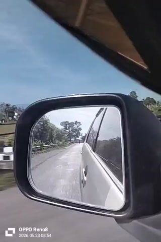 The video shows the view from a car side mirror while driving on a paved road. The mirror reflects the road behind the car, a motorcyclist, and trees lining the road. Fields and houses are visible in the distance. The sky is blue with some clouds. The side of the car is visible in the mirror as well.  The video displays a watermark for OPPO Reno8 and a date and time: 2026.05.23 08:54.