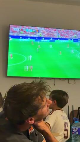 A group of people are watching a soccer game on a large television screen in a dimly lit room. A man with dark hair is in the foreground, leaning forward with his hand on his chin, intently watching the game. Behind him, a child wearing a white soccer jersey with the name "TRISTAN" and the number "5" on the back is also watching the game. A blue water bottle is visible on a table in the lower right corner of the frame. The television displays a soccer match with players in red and white uniforms on a green field. The score and time of the game are visible at the top of the screen. The environment appears to be a living room or common area.