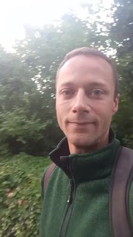 A man in a green fleece jacket and a backpack is standing in front of a background of trees. The camera is positioned at a slightly upward angle, showing the man's face in a close-up shot. The lighting is somewhat dim, suggesting it might be early morning or late evening. The man appears to be looking directly at the camera.