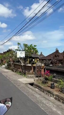 A point-of-view shot from a moving motorcycle, traveling down a straight, paved road in what appears to be a residential area. The sky is bright blue with scattered white clouds. Power lines stretch across the frame above the road. On either side of the road are traditional Balinese houses and stone walls covered in lush greenery. Other motorcycles pass by in the opposite direction. The mirror of the motorcycle taking the video is visible in the lower left corner of the frame.