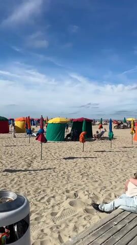 The video pans across a sandy beach on a sunny day. In the foreground, a couple is sitting on a wooden boardwalk, facing away from the camera. They are close together, with one person leaning their head on the other's shoulder. The beach is populated with numerous colorful umbrellas and beach tents, providing shade for people lounging. Further in the background, a boardwalk stretches along the beach, with more people walking and sitting. A large screen or billboard is visible on the boardwalk displaying images of people. The sky is blue with scattered white clouds. The overall scene depicts a lively and relaxed beach atmosphere.