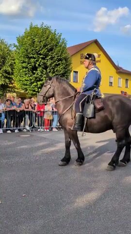A man on a black horse rides past a line of spectators. The man wears a blue jacket, dark pants, and a black hat with a white plume. He has a brown saddlebag on each side of the horse. The spectators are standing behind a metal barrier. A yellow building with a sign that says "Buttstädter Vollkornbäckerei" is in the background. The sky is blue with some clouds.