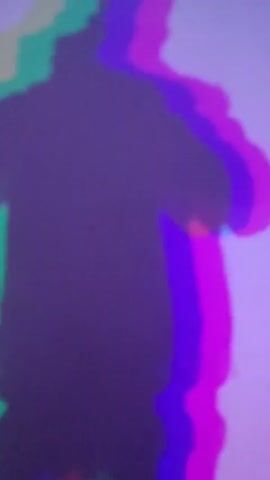 The media file presents a stylized shadow of a person cast against a pale, possibly lavender or off-white wall in an indoor setting. The shadow itself is dark and indistinct, but it is surrounded by vibrant, layered outlines in green, blue, and magenta, creating an effect reminiscent of an RGB split or chromatic aberration. The person's form is vague, appearing to be standing with one arm bent, possibly in a casual pose or mid-gesture.

The scene is dominated by this artistic lighting and shadow play. The presence of multiple colored outlines suggests that the shadow is created by either multiple colored light sources or a single light source passing through a prism or filter. No specific activity beyond posing for the shadow effect is evident.

Notable details include the striking visual effect of the multicolored shadow, which gives the image a modern, experimental, or glitch-art aesthetic. The background wall is otherwise plain, serving as a canvas for the shadow. Artificial lighting is clearly in use to create this specific visual. There are no indications of time of day or weather, nor are there any discernible location cues within the frame itself. No text is visible.