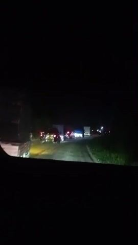 The video shows a nighttime traffic jam on a road. Headlights from various vehicles illuminate the scene. There are several cars and trucks visible, including a few motorcycles. The road appears to be unpaved in some areas. There is some vegetation visible on the right side of the road. The lighting is dim, and the overall atmosphere is dark. The vehicles are stationary, indicating a traffic standstill.