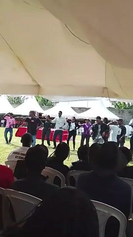 The video shows a group of about ten people dancing in unison on a slightly raised platform, likely a stage, under a large white tent. Most of the dancers are wearing casual clothing, like jeans, t-shirts, and jackets. One person is wearing a white dress shirt. A small crowd of people is seated in chairs facing the stage, watching the performance.  The tent structure suggests an outdoor event.  There are more white tents visible in the background. The grass is green, and there are some trees and a stone wall in the distance. A red carpet covers the stage.