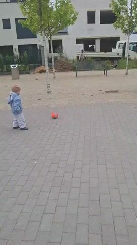 A man in a white tracksuit stands in a park and watches as a toddler wearing a blue jacket and a tan hat plays with an orange ball. The toddler crouches, picks up the ball, and then stands and begins to kick it around. The park has a paved walkway, gravel, trees, benches, and construction in the background.
