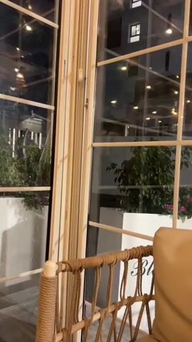 A camera pans across a window, showing a view of a balcony at night. There are string lights visible on the balcony, and several plants. A wicker chair with a tan cushion is in the foreground.
