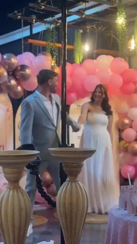 A man in a light blue suit and a woman in a white dress are standing next to each other in front of a backdrop of pink and gold balloons. The man is looking at the woman and smiling, while the woman is looking at the man and smiling. There are two decorative pillars in the foreground of the image, one on the left and one on the right. The scene appears to be a celebration or event, possibly a wedding or engagement party, given the attire and decorations. The background also includes some text in Hebrew, which is partially visible. The lighting appears to be artificial and somewhat dim.