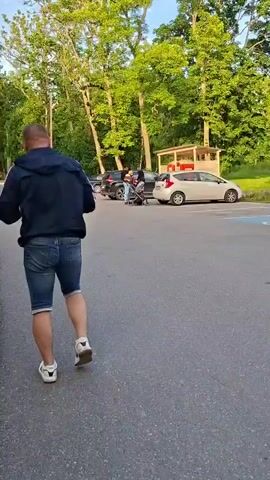 A man wearing a blue jacket and denim shorts walks away from the camera on a paved path. He is walking towards a parking lot with several cars and trees. In the background, a couple with a baby stroller is visible near one of the cars. The scene appears to be outdoors during the daytime.