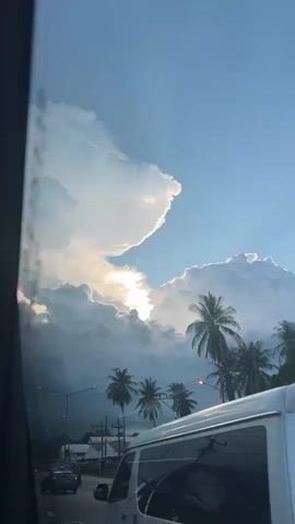 The video captures a dramatic sky over Tambon Don Sak, Thailand, likely during the late afternoon or early evening. Towering cumulonimbus clouds dominate the frame, with the sun breaking through a gap, creating a bright, almost blinding light that illuminates the edges of the clouds. A faint rainbow effect is visible where the light interacts with the moisture in the clouds.

In the foreground, a road is visible, with a few cars and motorcycles driving on it, suggesting this is a public thoroughfare. Silhouettes of palm trees line the road, a clear indicator of a tropical or subtropical location. Streetlights are also present, one with a distinctive double arm, further contributing to the scene's setting. The overall mood is one of awe at the power and beauty of nature, with the dramatic sky contrasting with the ordinary, everyday activity on the road below. The weather appears to be transitioning, possibly after a rain shower, given the intense cloud formations and the presence of the rainbow effect. There is no visible text in the video.