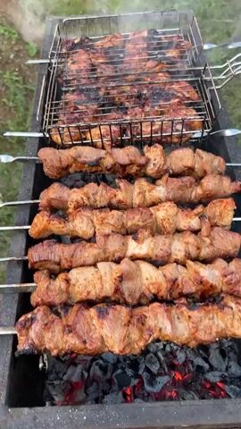The video shows a close-up, overhead view of a barbecue grill with various meats cooking over hot coals. The scene is set outdoors, with green grass and foliage visible in the background, suggesting a natural or semi-rural setting. The lighting indicates it is daytime, possibly late afternoon or early evening, with the sun casting a soft glow.

The grill contains several skewers of meat, appearing to be pork or lamb, arranged in rows. The meat is well-marinated and has a slightly charred, caramelized appearance, indicating it is nearing completion. Above the skewers, a metal grill basket holds pieces of chicken or ribs, also visibly cooking and developing a browned exterior. Wisps of smoke rise from the grill, carrying the aroma of the cooking meat.

The activity is the preparation and cooking of a barbecue. The focus is entirely on the food and the process of grilling. There are no people visible in the frame, and no text is present. The overall impression is one of delicious food being prepared for an outdoor gathering, evoking feelings of warmth, satisfaction, and anticipation of a shared meal. The image captures a moment of culinary enjoyment in a natural outdoor environment.