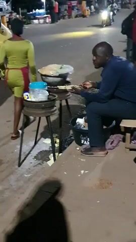 A man sits on a stool in front of a street food stall. He is wearing a dark blue shirt and dark pants. In front of him is a large pot on a stand with a charcoal brazier. He appears to be eating something with his hands. A woman in a green dress and red apron walks behind the stall. The stall is set up on a sidewalk next to a street. There are other people and vehicles in the background, and the scene appears to be at night.