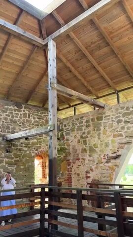 A low-angle shot captures a woman in a white outfit standing in the foreground, looking up. She is positioned behind a wooden railing. The background reveals a stone wall with an arched opening and rough-hewn stones. Above, a wooden roof structure is visible, with rafters made of natural wood beams supporting a wooden plank ceiling. A large, open rectangular section in the roof allows a view of a bright blue sky filled with scattered white clouds. The overall scene suggests the interior of an old, partially renovated structure with an exposed roof and stone walls, possibly a historical ruin or an ancient building.