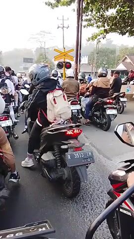 The video shows a traffic jam of motorcycles stopped at a railway crossing. The perspective is from a motorcycle behind other motorcycles. Several riders are wearing helmets and backpacks. A traffic signal with red and yellow lights is visible, along with a railway crossing sign. The road appears to be paved and wet in some areas. The sky is overcast or hazy. The license plates of some motorcycles are visible, showing registration numbers. The scene suggests a rural or suburban setting.