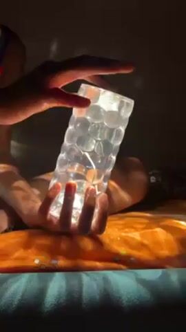 A person sitting on a bed with an orange patterned bedspread is holding a cylindrical lamp with a clear, textured surface resembling bubbles. The lamp is emitting a bright white light, casting shadows and illuminating the person's hands. The room appears to be dimly lit, with a strong beam of light shining from the side, adding to the contrast. One hand is holding the lamp from the bottom, and the other hand is placed on top, partially covering the lamp.