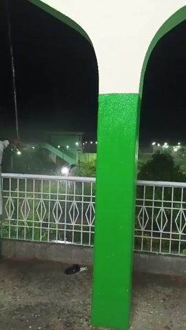 The video shows a night scene with a green and white arched structure in the foreground. Beyond the structure, there is a white metal fence and trees. In the background, there are lights and buildings, suggesting an outdoor area or a park. Some water is visible, possibly indicating rain or sprinklers. There is a black object on the ground near the fence, which appears to be a shoe or sandal. A person is partially visible on the far left, holding a hose and spraying water. The overall atmosphere is dark and somewhat wet.
