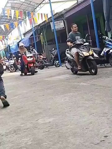 In a wide shot from a low angle, a street scene is captured with several motorcycles parked and in motion. The ground is paved with rough asphalt, and a covered walkway with colorful triangular flags strung across it stretches along the right side of the street. Several shops with open fronts are visible beneath the walkway. On the left side, a person on a red scooter is facing the camera, wearing a light-colored headscarf and a long-sleeved shirt. Behind them, a person on a pink scooter is moving away from the camera, wearing a light-colored headscarf and a patterned dress. Further down the street, other motorcycles and people are visible, with one man on a black motorcycle in the foreground, to the right, wearing a light-colored shirt and dark shorts, smiling as he rides away from the camera. The overall scene suggests a market or a busy street in a tropical or subtropical region.
