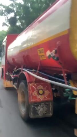 This media file captures a close-up, slightly shaky video of a red and yellow water tanker truck driving on a paved road. The truck has a large cylindrical tank, with a white stripe running horizontally along its length. The rear of the truck features a yellow ladder, and decorative floral paintings in red, yellow, and green on the mud flaps. A visible "STOP" sign is also present on the back. Text visible on the truck includes "TW03 F-9711" and what appears to be "Soun...", possibly part of a company name. The Indian flag is also depicted on the side of the tanker. The scene appears to be set during the day, possibly late afternoon, given the lighting. Trees and buildings line the background, suggesting an urban or suburban environment. The weather seems overcast. The video is filmed from a moving vehicle, likely another car, indicating the tanker is in transit.