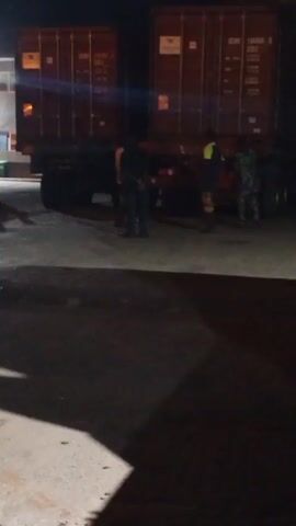 The video shows a dimly lit outdoor area at night, seemingly a loading or storage facility.  Several people are present, some standing near a large truck with two cargo containers on its trailer.  The figures are silhouetted, making individual identification difficult.  A white building is visible in the background, illuminated by an overhead light.  The ground is concrete and appears wet in places, casting reflections.  The overall impression is one of activity around the truck, possibly related to loading or unloading cargo.