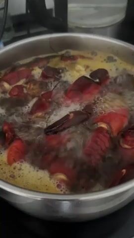 A close-up shot shows a pot of boiling water on a stove filled with crayfish. The crayfish are bright red and appear to be cooked. Bubbles are rising to the surface of the water, indicating that it is at a rolling boil. The pot is made of stainless steel and has a silver rim. The stove top is black and appears to be an electric coil stove. The background is slightly blurred but appears to be a kitchen counter with some other kitchen items visible. The overall impression is one of preparing food, specifically cooking crayfish.