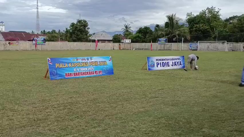 The video shows a football field with a soccer game underway. The field is grassy and well-maintained. In the background, there are buildings and trees. A group of people are gathered near the field, some of whom are wearing blue shirts with a logo that says "PEMERINTAH KABUPATEN PIDIE JAYA." A young boy is walking on the field, holding a soccer ball. There is a trophy on display, which appears to be part of the event. The overall atmosphere seems to be one of a community event or sporting competition.