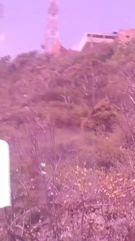 The video shows a view from a moving vehicle, likely a car, looking up a hillside. The hillside is covered in sparse vegetation and rocks. Dominating the top of the hill are several tall radio towers and some buildings, suggesting a communications hub or a research facility. The sky is a hazy, pale purple, indicating either dawn, dusk, or possibly an unusual atmospheric condition. The lighting suggests it is daytime, but the color cast gives a dreamlike or surreal quality to the scene.

There are no people visible in the video. The objects in focus are the radio towers, buildings, and the general landscape of the hillside. The scene is an outdoor, hilly environment with man-made structures on an elevated position.

The activity taking place is motion; the perspective is clearly from a moving vehicle, indicated by the slight blur and the changing view of the landscape. The time of day is suggested to be daytime, but the muted and purplish hue of the light is a notable detail, creating an unusual atmosphere. The location cues are the hilly terrain and the presence of multiple communication towers, characteristic of elevated sites used for signal transmission. There is no discernible text in the video.
