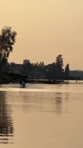 The video shows a person riding a jet ski on a calm lake at dusk. The sky is a pale yellow-orange, and the water reflects the color of the sky. The jet skier is in the center of the frame, moving towards the viewer, leaving a wake behind. Along the shoreline on the left and right are houses, trees, and docks. There is also a person on a kayak or paddleboard in the distance. The overall scene is tranquil and peaceful.