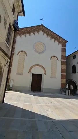 A vertical video pans across a town square on a sunny day. The first shot shows the facade of a church with a cross on top, arched windows with decorative elements, and a large wooden door. The second shot pans to the right, revealing more buildings with arched walkways and windows. The third shot continues the pan, showing a building with the word "MUNICIPIO" (municipality) on it. The final shots focus on a tall bell tower and a building with arches and flags displayed on a balcony. The square is paved with light-colored stone tiles. There are no people visible in the video.