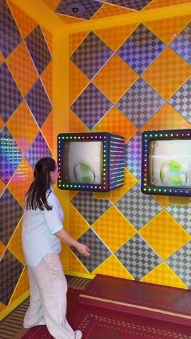 A woman enthusiastically interacts with three Super Mario-themed display blocks in an indoor setting. She wears a light blue short-sleeved top and light-colored baggy pants. The room features walls and a ceiling adorned with a vibrant checkerboard pattern of orange, yellow, and grey/purple squares, characteristic of the Super Mario universe. The three square blocks are mounted on the wall at eye level and are surrounded by colorful, flashing LED lights.

The woman repeatedly jumps and hits the bottom of these blocks with her hand, mimicking how Mario interacts with "question blocks" in the games. Each hit causes the image displayed on the block's screen to change. Visible power-ups include a yellow star, a fire flower, and a green Yoshi egg. She is clearly enjoying the activity, smiling and laughing throughout the video. The floor is covered with a red carpet featuring a gold trim at the base of the wall. The scene suggests an interactive exhibit or game within a theme park, likely Super Nintendo World at Universal Studios Japan in Osaka. No visible text is present.