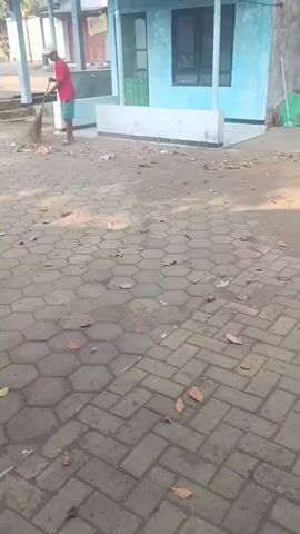 A person wearing a red shirt and a white hat is sweeping the ground with a broom. The person is standing on a paved area with hexagonal tiles. There is some debris on the ground, including leaves and trash. In the background, there is a building with a blue wall and a white structure.