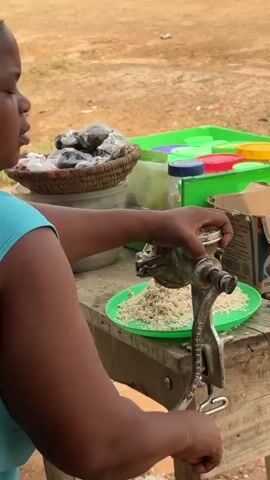 A woman with dark skin and braided hair, wearing a teal tank top, is operating a manual grain grinder. The grinder, made of metal, is clamped to a wooden table. A green plate filled with finely ground grain sits beneath the grinder's output spout. In the background, a woven basket holds several dark, irregularly shaped objects, possibly dried food items, and plastic containers are visible. The ground is dusty and reddish-brown, suggesting an outdoor setting. The woman's right hand is on the grinder's crank, and her left hand is holding the device steady. She is positioned to the left of the grinder, facing away from the viewer. The overall impression is of food preparation in a rural or market environment.