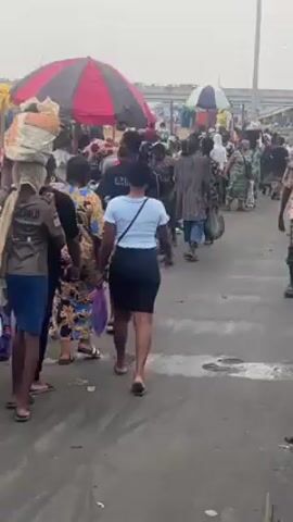 The video shows a crowded outdoor market with many people walking in different directions. Some people are carrying goods on their heads. There is a large red and black striped tent in the background. The ground appears to be wet. The sky is overcast. The clothing of the people varies, with some wearing casual attire and others wearing more traditional African fabrics.