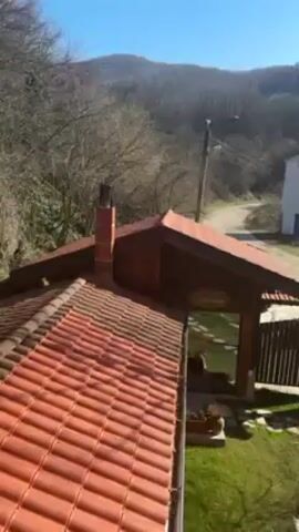 The video shows a landscape from a slightly elevated viewpoint, possibly a balcony or upper floor of a building. In the foreground, there's a red-tiled roof of a small structure, possibly a gazebo or a small building extension, with a chimney visible on the left side of the frame. Beyond the roof, a winding road leads away from the camera, and a small white building can be seen alongside the road. The background consists of a forested hillside under a clear blue sky. As the video continues, the camera pans slightly to the right, revealing a birch tree, a parking area with a couple of vehicles, and a Bulgarian flag hanging from the balcony railing.