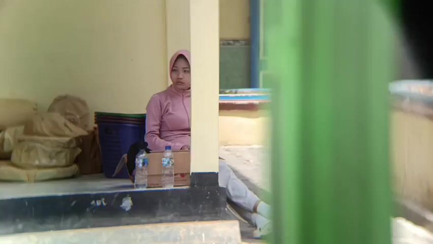 A young woman wearing a pink hijab and pink tracksuit is sitting on a step, looking down. To her left, there are several bags and a blue plastic basket. In front of her, there are two bottles of water and a brown box. The background shows a building with light-colored walls and some colorful decorations. The scene appears to be outdoors, possibly on a porch or under an awning. A blurry green object is in the foreground, obscuring part of the view.