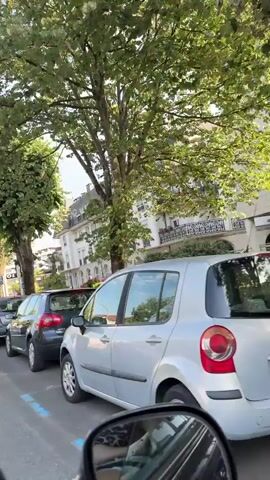 The video shows a street view from a car. In the foreground, there is a car's side mirror. The middle ground features several parked cars, including a dark blue Volkswagen Golf with a license plate starting with "3447 FKC," and a grey Fiat 500. Behind the cars, a multi-story building with a distinctive mansard roof and balconies is visible. Trees with green foliage line the street, and a traffic light is also present. In the background, a person is walking away from the camera on a sidewalk. Two large trash bins are situated near the foreground. The scene appears to be a street in a European town, possibly during daytime.
