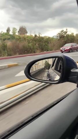 The view from a moving car shows the side mirror. The mirror reflects the road behind the car, with other cars and buildings visible. A sign is visible that reads, "Objects in mirror are closer than they appear." A red car is seen in the background, traveling in the same direction. The weather appears overcast.