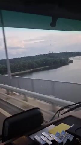 The video, filmed from the dashboard perspective of a moving vehicle, captures a drive across a long bridge during dusk or early evening. The initial view from inside the vehicle shows the windshield, a part of the dashboard, and a device mounted near the rearview mirror. Outside, the bridge's guardrail is visible, stretching alongside a wide body of water, likely a river. Beyond the water, a distant landmass covered with trees can be seen under a soft, cloudy sky.

As the vehicle proceeds, the camera pans to show the road directly ahead. The bridge features a multi-lane asphalt roadway with clear white lane markings. White guardrails line both sides of the bridge. Numerous streetlights extend into the distance, some already illuminated, casting a soft glow. A distinctive feature on the left side of the bridge structure is the presence of overhead lines and supports, characteristic of a railway track running parallel to the vehicular road. The sky remains overcast or partly cloudy, indicating a quiet transition from day to night. The road ahead appears relatively clear of other vehicles. The scene depicts a calm journey across a significant infrastructure overpass, consistent with a bridge crossing a river in a location like Antimovo, Bulgaria. No people or legible text are visible within the frame.