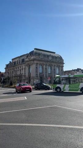 A busy street scene unfolds under a clear blue sky. Dominating the background is a large, ornate, classical-style building with an arched roof. In the foreground, a multi-lane intersection is active with traffic. A red SUV is turning left, a black car is visible behind it, and a white compact car is driving straight. To the right, a green and white bus with the text "BALADO 100% electric" is stopped, likely waiting for traffic or passengers. The road surface is asphalt, with clear lane markings. The overall impression is of a typical urban environment on a bright, sunny day.